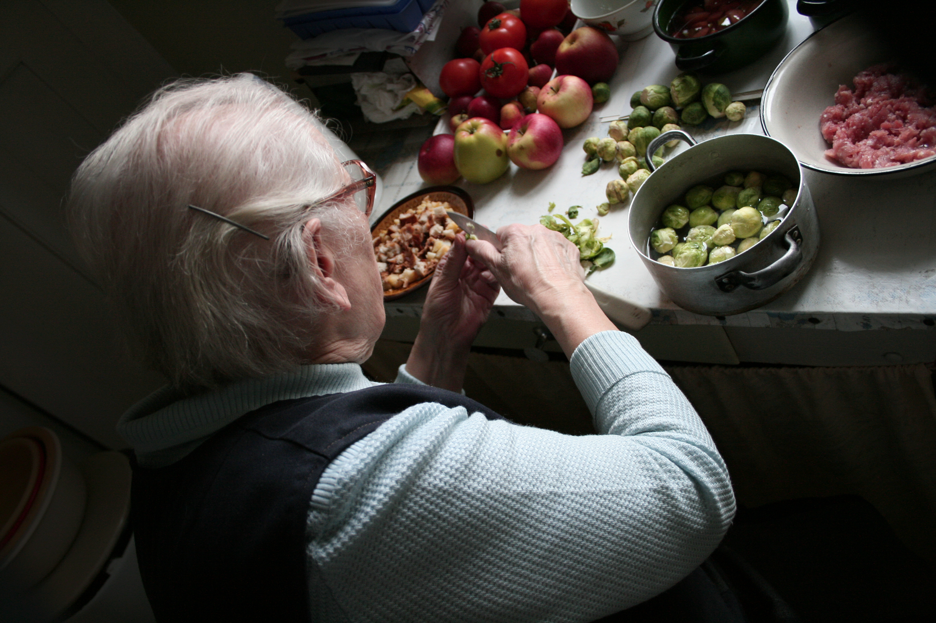 An elderly woman peels vegetables in a cozy kitchen setting, surrounded by fresh produce.