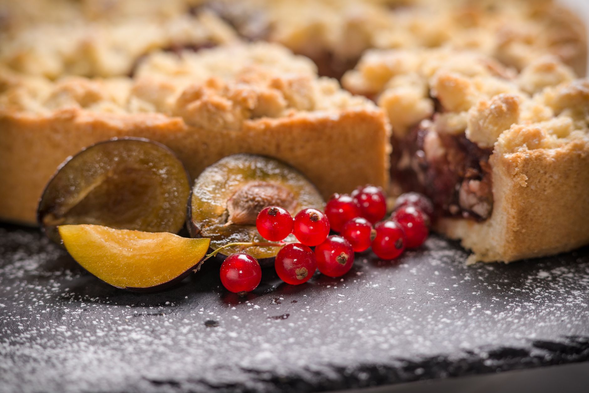 Close-up of a plum crumble tart with red currants, dusted with powdered sugar.