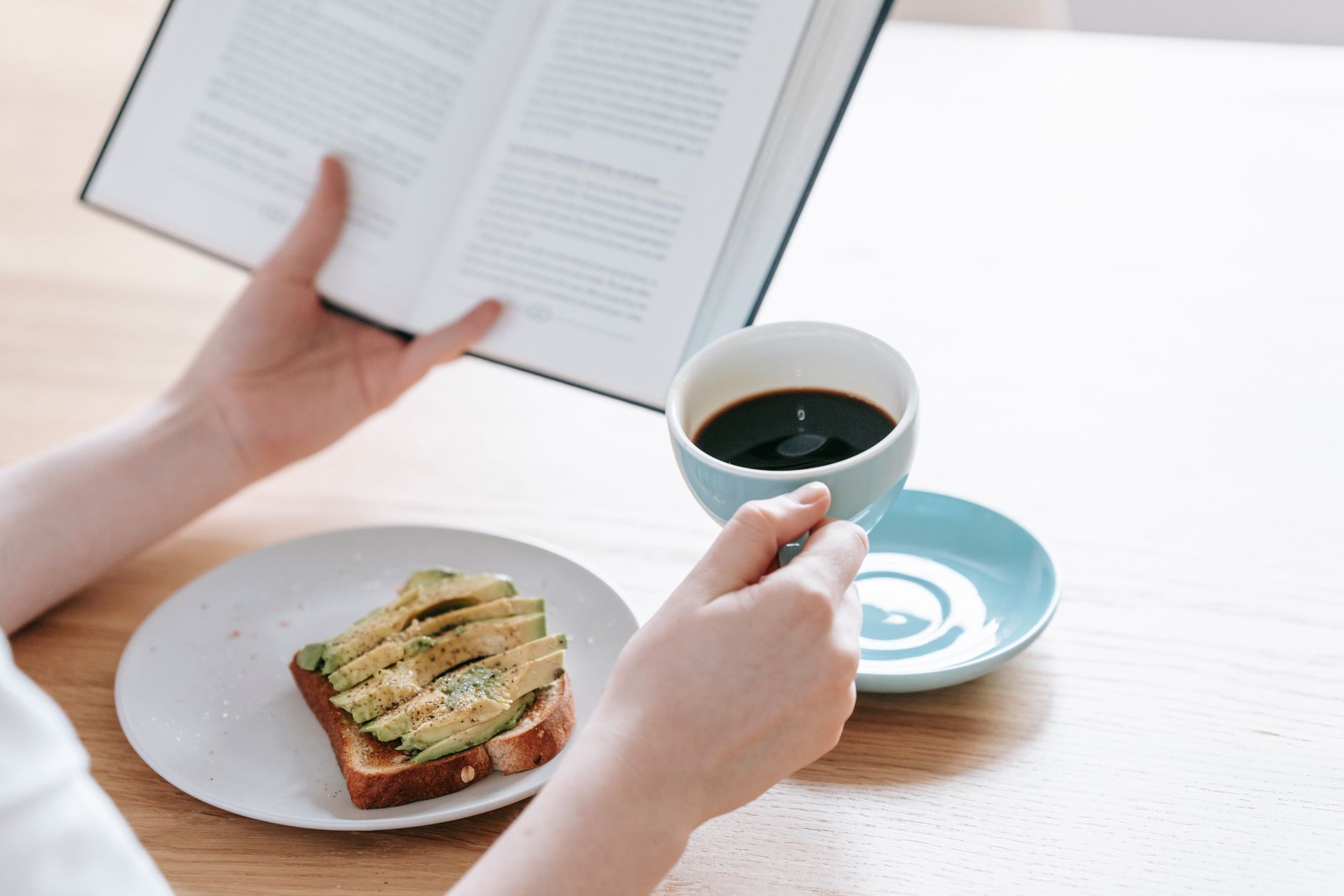 Faceless female sitting at table with cup of fresh black coffee and avocado toast having breakfast and reading book