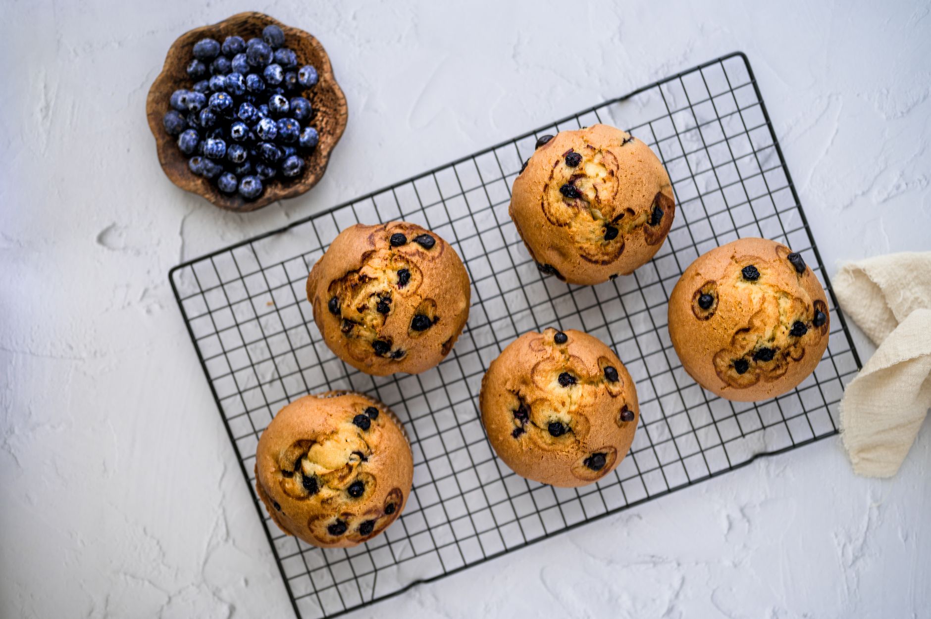 Top view of homemade blueberry muffins cooling on a wire rack with fresh blueberries in a bowl.