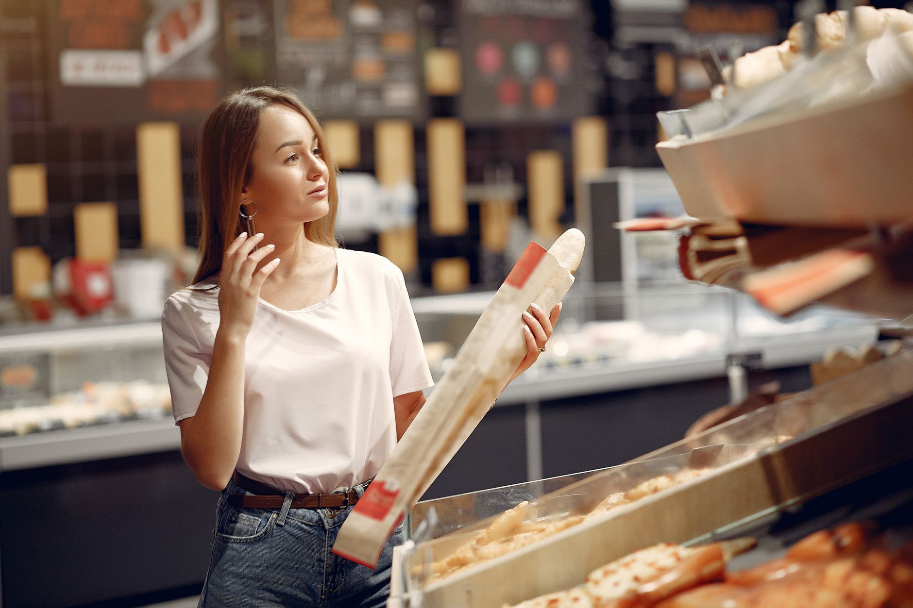 Happy young woman with long hair in casual outfit taking baguette  and choosing yummy bun on showcase while visiting supermarket