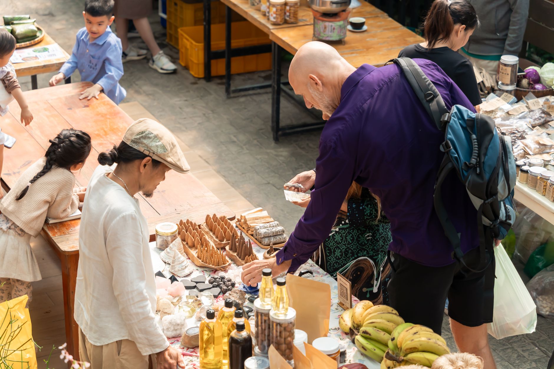 A vibrant outdoor market with diverse people browsing and buying assorted products from vendors.