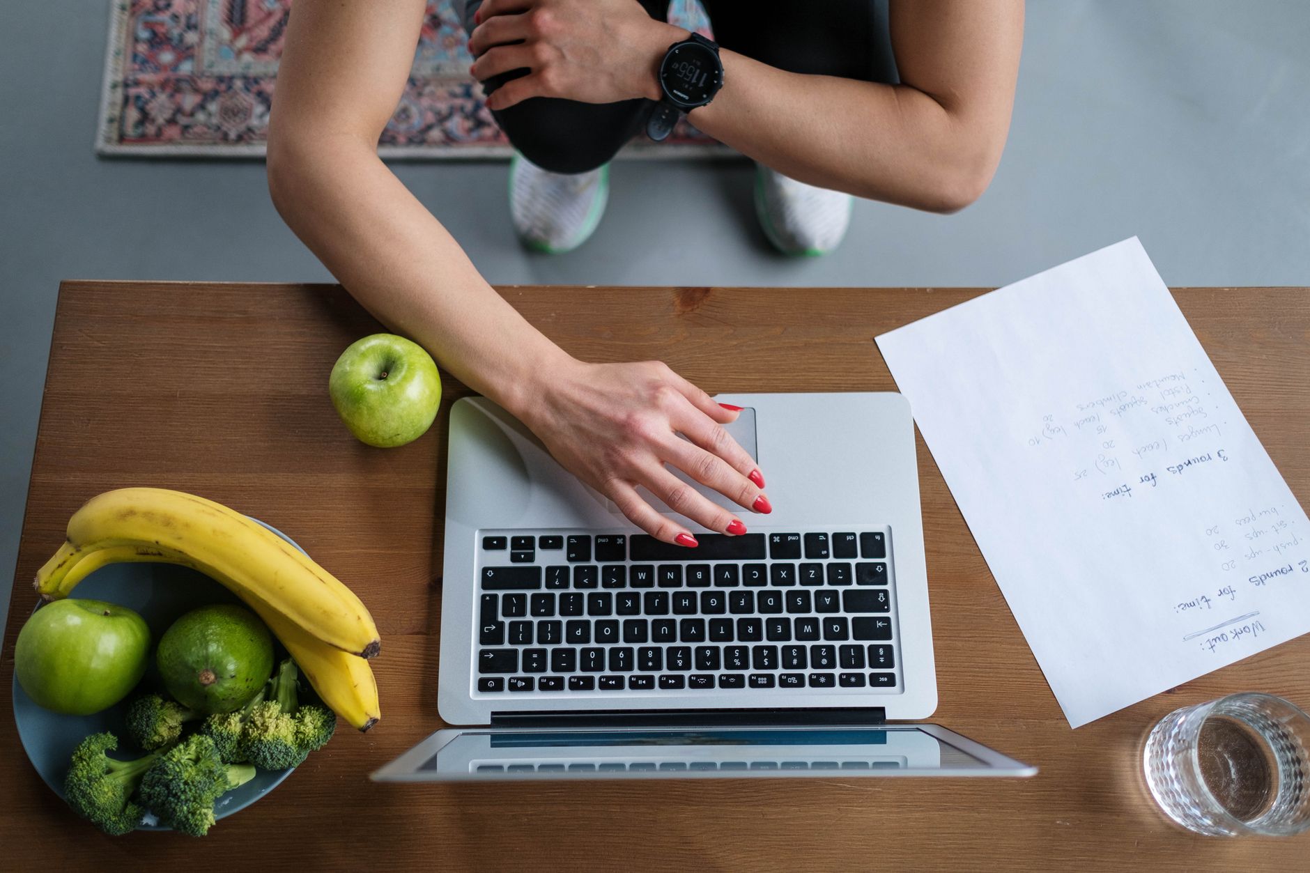 Overhead view of hands on laptop with fruits, vegetables, and paper on wooden table.