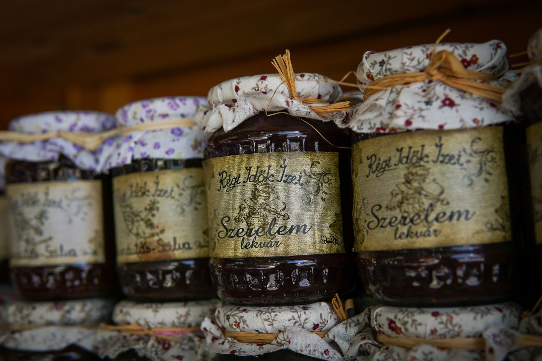 Close-up of rustic homemade jam jars with floral fabric covers and rustic labels on a wooden shelf.