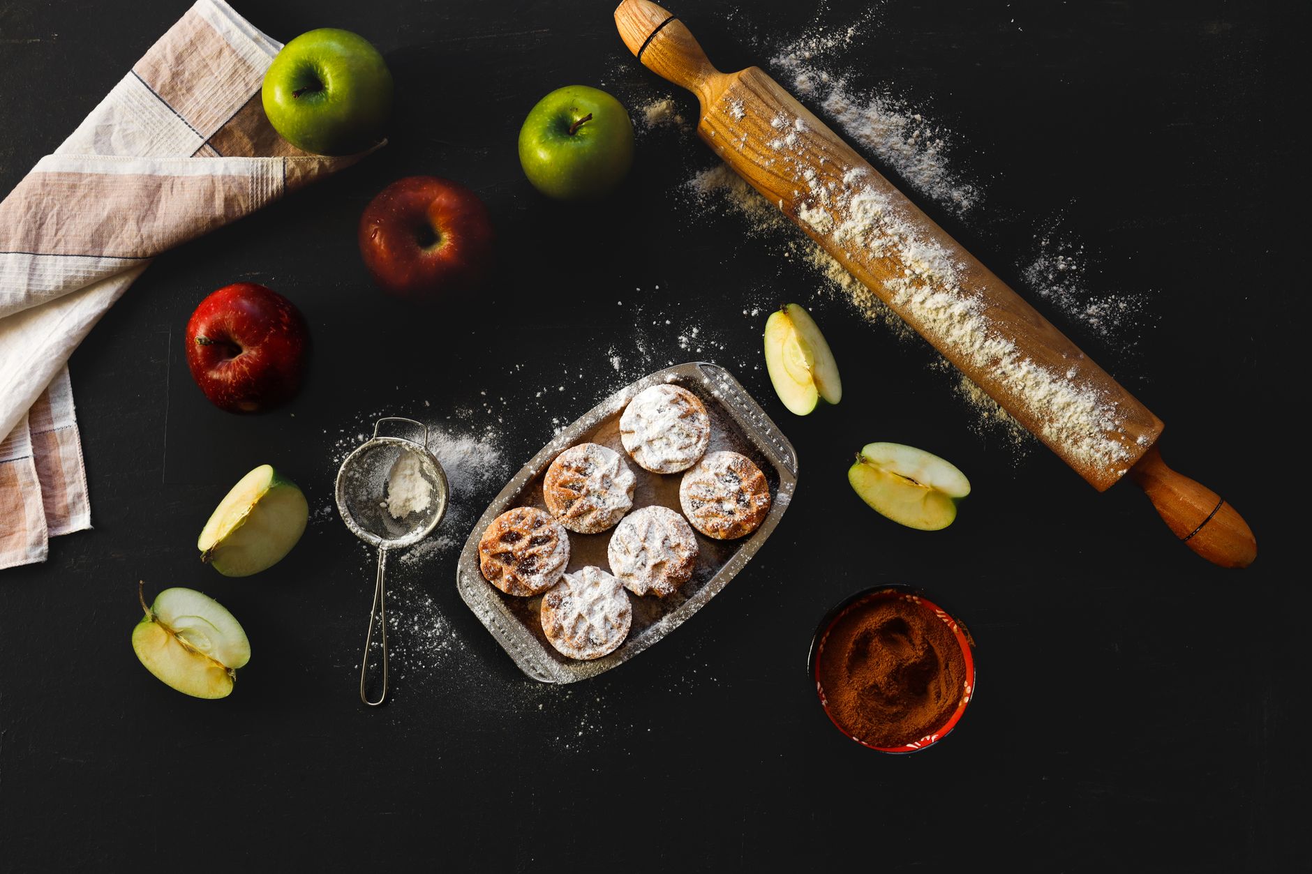 Flat lay of apple pie preparation with fresh apples, rolling pin, and pastry on dark surface.