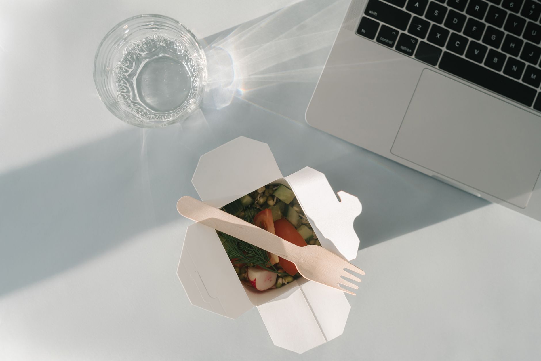 Healthy salad in a takeout box with a glass of water and a laptop on a white surface, top view.
