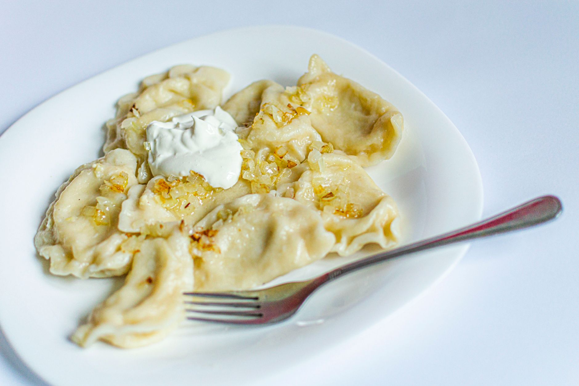 Plate of handmade dumplings with caramelized onions and sour cream on a white background.
