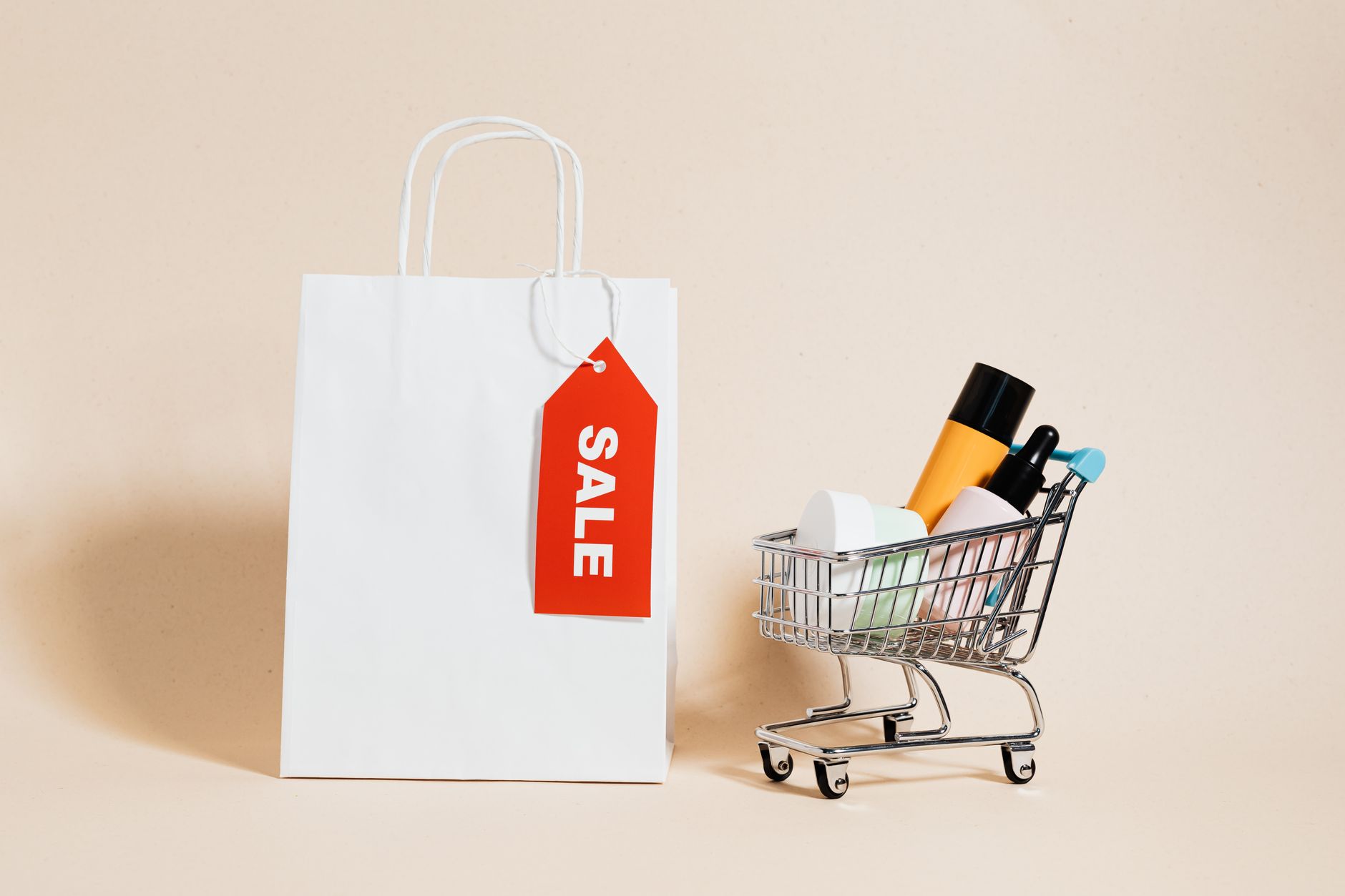 White shopping bag with red sale tag and mini shopping cart on beige background.