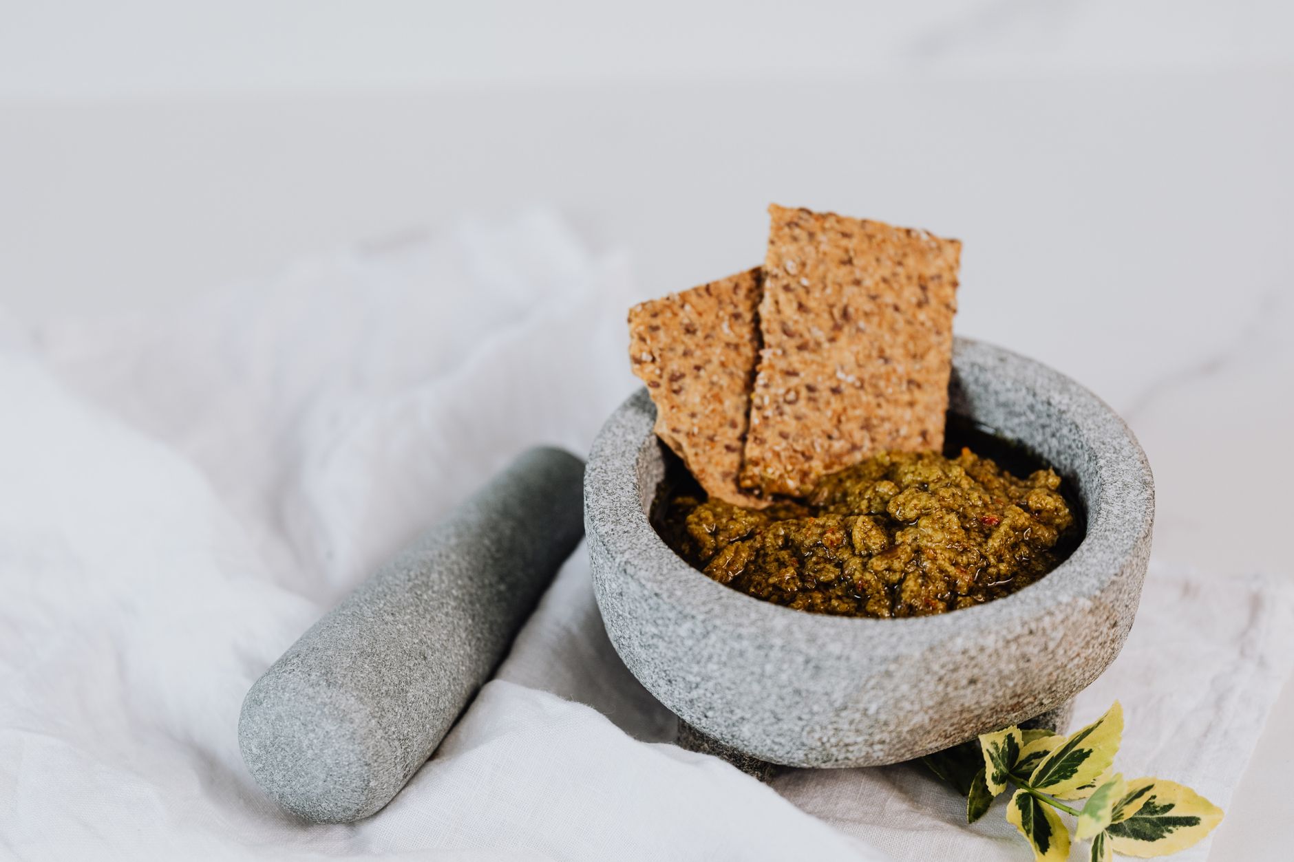 Stone mortar and pestle with fresh green pesto and crispbreads on white cloth.