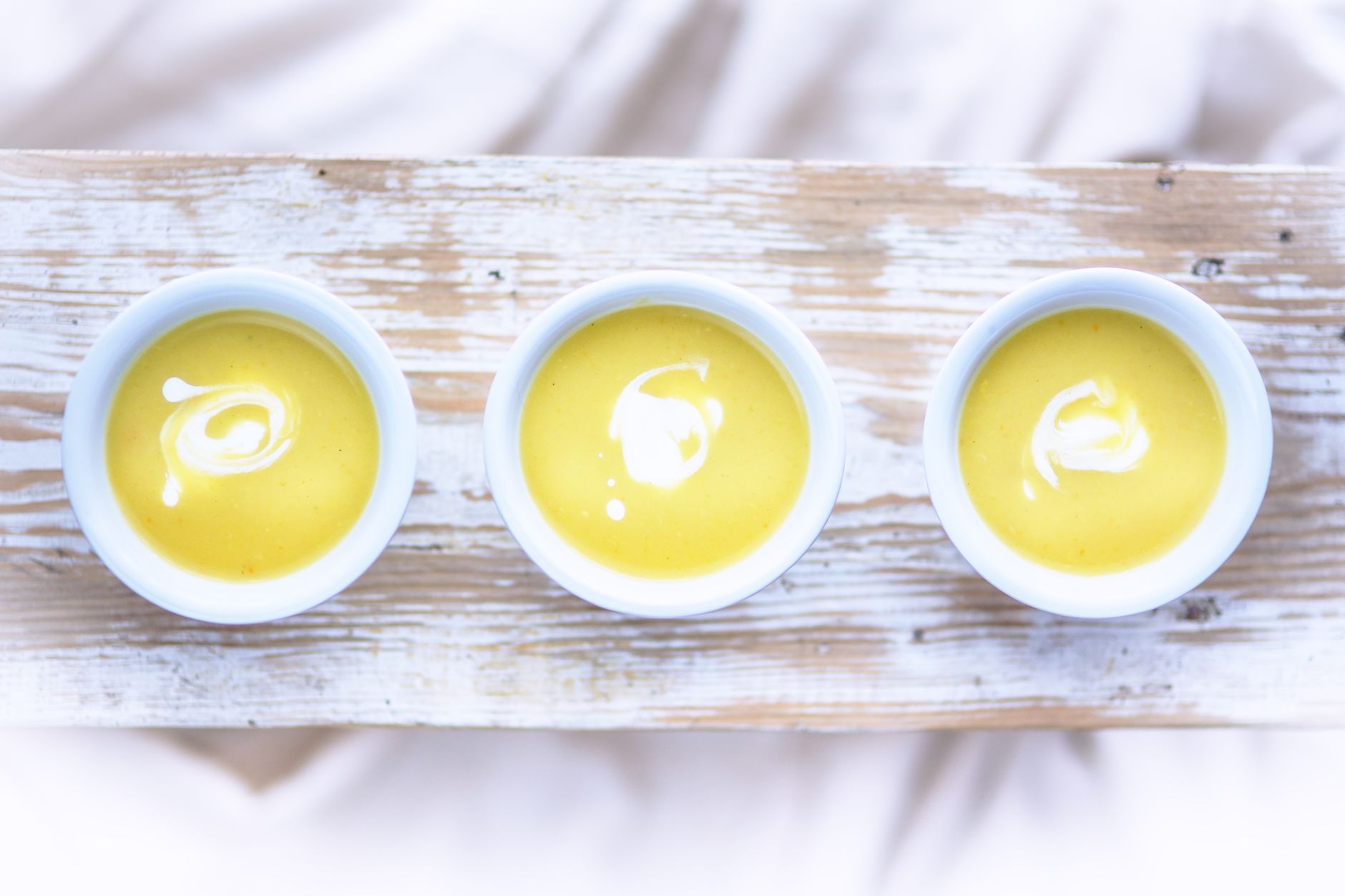 Top view of three bowls of creamy yellow soup with drizzle, on a rustic wooden table.