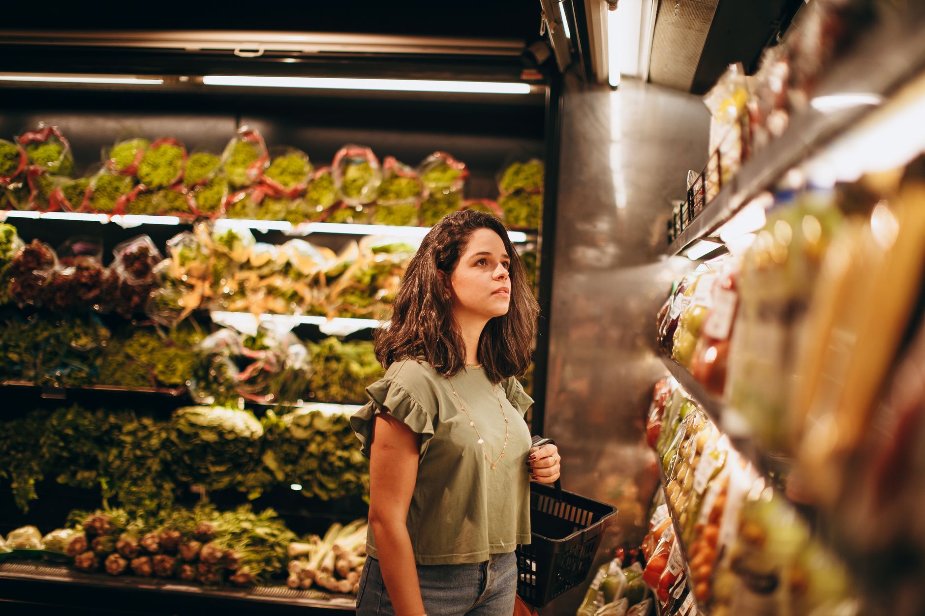 Woman browsing fresh produce with basket in supermarket. Indoor shopping scene.
